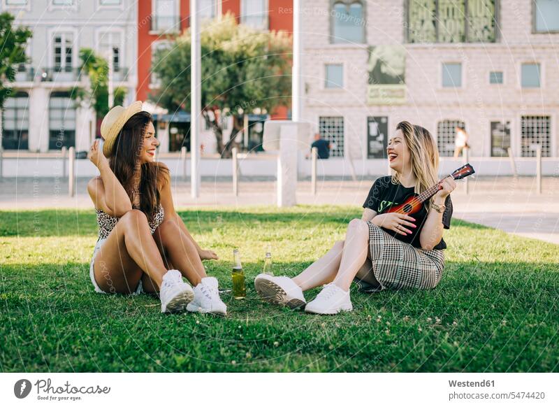 Cheerful woman playing ukulele while sitting with female friend on grassy land color image colour image Portugal leisure activity leisure activities free time
