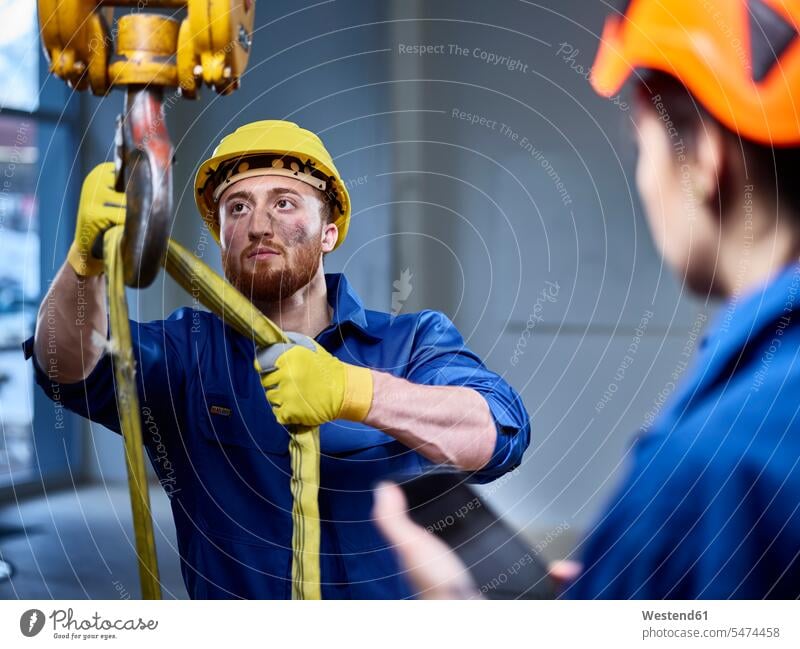 Industrial worker fixing hoist sling on indoor crane, female colleague controlling with digital tablet accessibility accessible hook hooks overall overalls