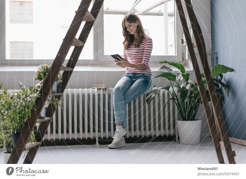 Woman sitting on radiator in her new home, reading e-book, surrounded by plants woman females women E-Book ebook electronic book at home potted plant