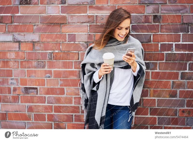 Happy woman with coffee to go looking at cell phone in front of brick wall Coffee to Go takeaway coffee brick walls Smartphone iPhone Smartphones females women