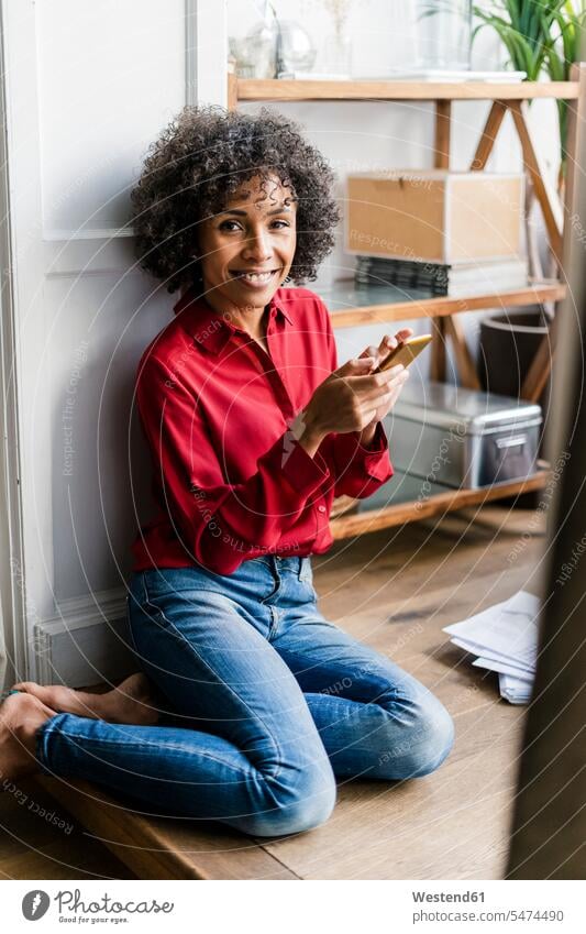 Portrait of smiling woman sitting on the floor at home with cell phone portrait portraits females women floors Seated smile mobile phone mobiles mobile phones
