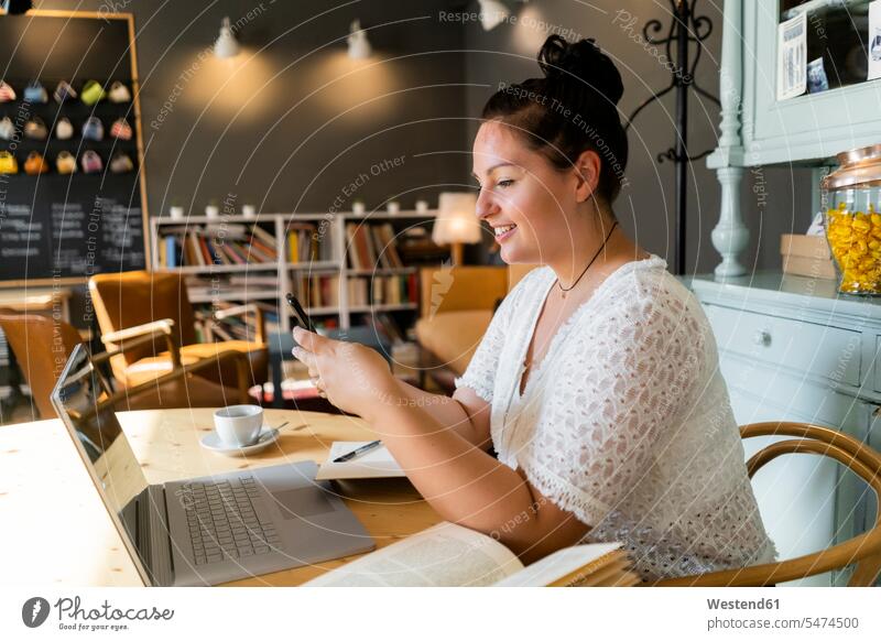 Young woman with books and laptop on table using smart phone in coffee shop color image colour image indoors indoor shot indoor shots interior interior view