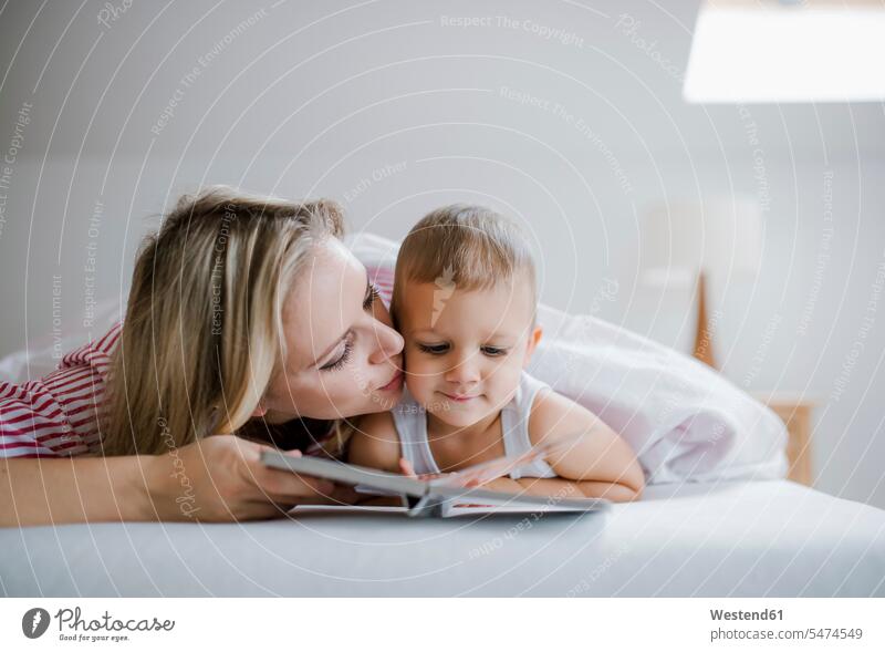 Mother kissing toddler son lying in bed at home reading a book books laying down lie lying down mother mommy mothers ma mummy mama sons manchild manchildren