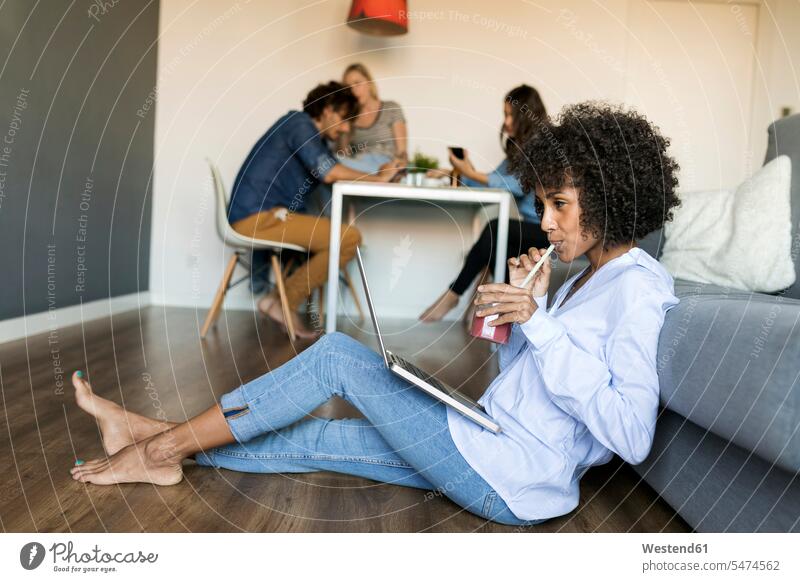 Woman with soft drink sitting on floor using laptop with friends in background floors woman females women refreshing drink soft drinks refreshing drinks Seated