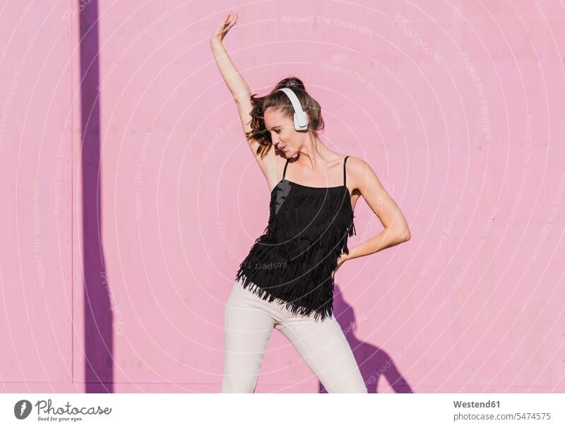 Happy young woman wearing headphones dancing in front of pink wall headset happiness happy dance females women Rosy walls Adults grown-ups grownups adult people