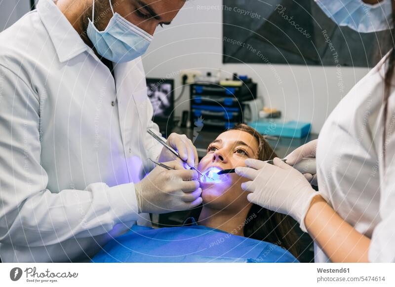 Male dentist in mask and gloves examining teeth of female patient with help of assistant at clinic color image colour image indoors indoor shot indoor shots