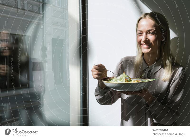Smiling young woman standing at the window in sunshine eating a salad blond blond hair blonde hair healthy eating nutrition dieting health awareness