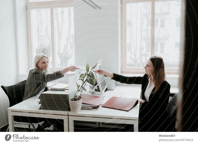 Two young women working at desk in office handing over paper filing tray sitting Seated Cooperation working together collaboration Cooperating Cooperate