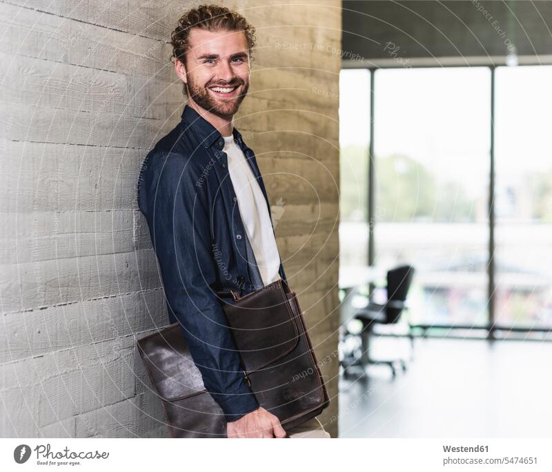Smiling casual businessman standing on office floor holding briefcase offices office room office rooms portrait portraits men males office suite brief case
