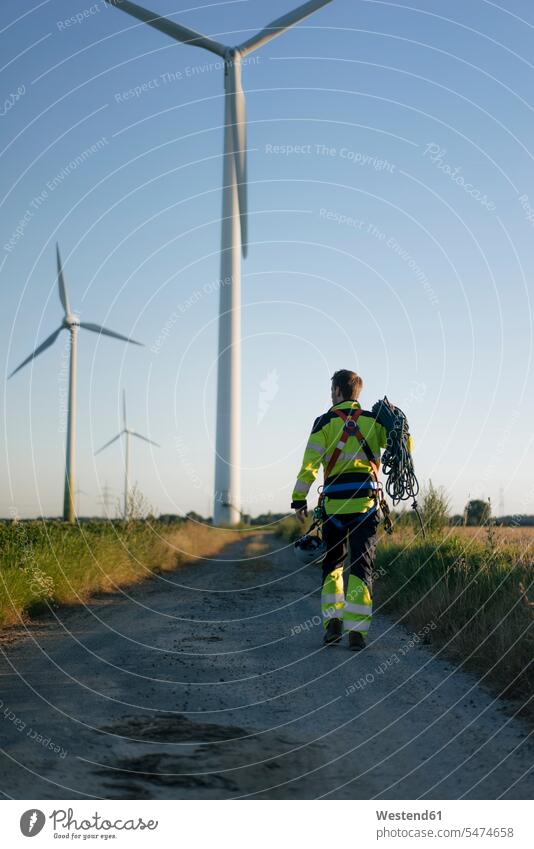 Technician walking on field path at a wind farm with climbing equipment going country lane field road wind park technician technicians wind power plant