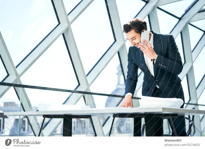 Smiling businessman on cell phone looking at plan in office eyeing on the phone call telephoning On The Telephone calling plans Businessman Business man