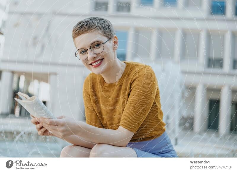 Smiling mid adult woman with book sitting against fountain color image colour image outdoors location shots outdoor shot outdoor shots day daylight shot