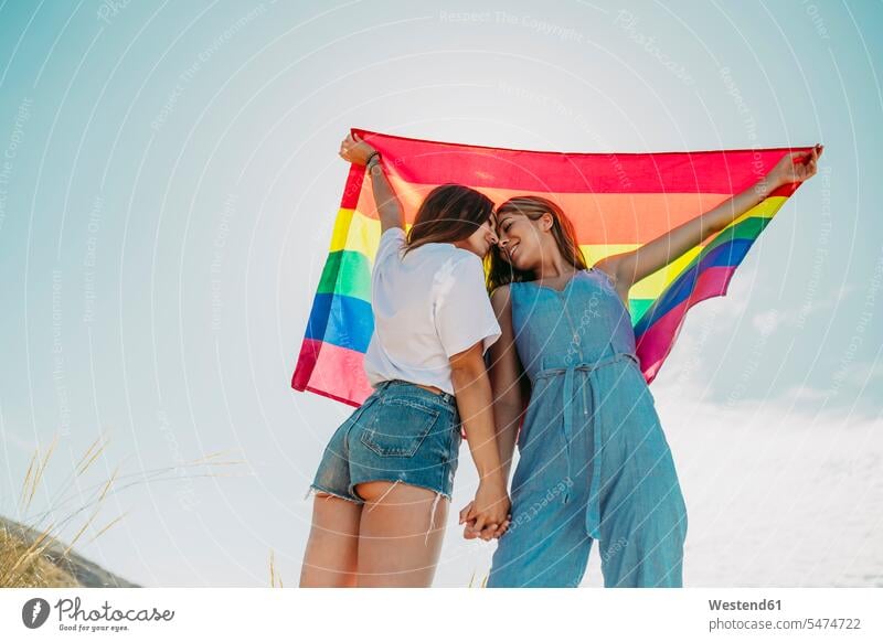 Two affectionate young women holding an LGBT flag under blue sky (value=0) friends mate female friend Female Friendship human human being human beings humans