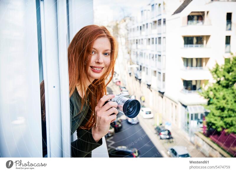Portrait of smiling redheaded woman with camera leaning out of window cameras portrait portraits females women windows red hair red hairs red-haired Adults