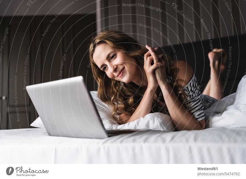 Smiling woman lying on bed at home looking at laptop Laptop Computers laptops notebook laying down lie lying down beds females women eyeing computer computers