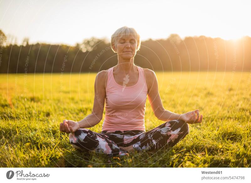 Senior woman in lotus position on rural meadow at sunset country countryside sunsets sundown females women senior women elder women elder woman old senior woman