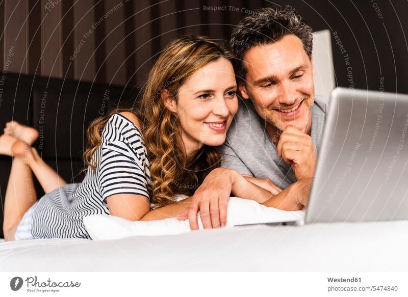 Happy couple lying on bed at home looking at laptop twosomes partnership couples laying down lie lying down beds eyeing happiness happy Laptop Computers laptops