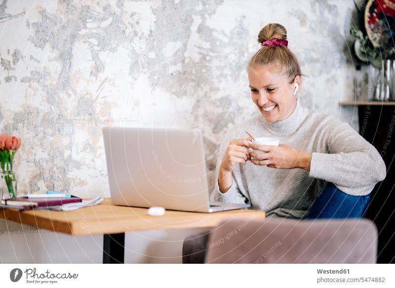 Smiling blond woman sitting on armchair at home having video chat with laptop and earphones human human being human beings humans person persons