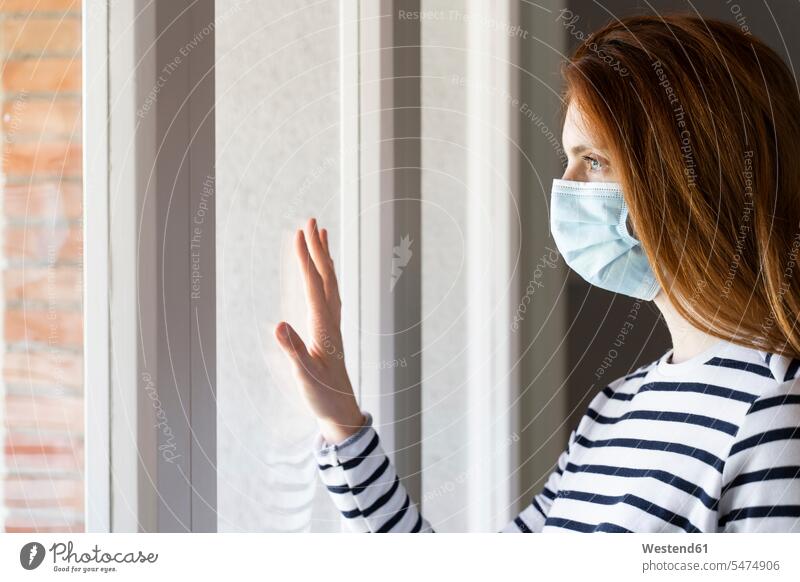 Redhead woman wearing protective face mask standing by window at home during COVID-19 color image colour image indoors indoor shot indoor shots interior