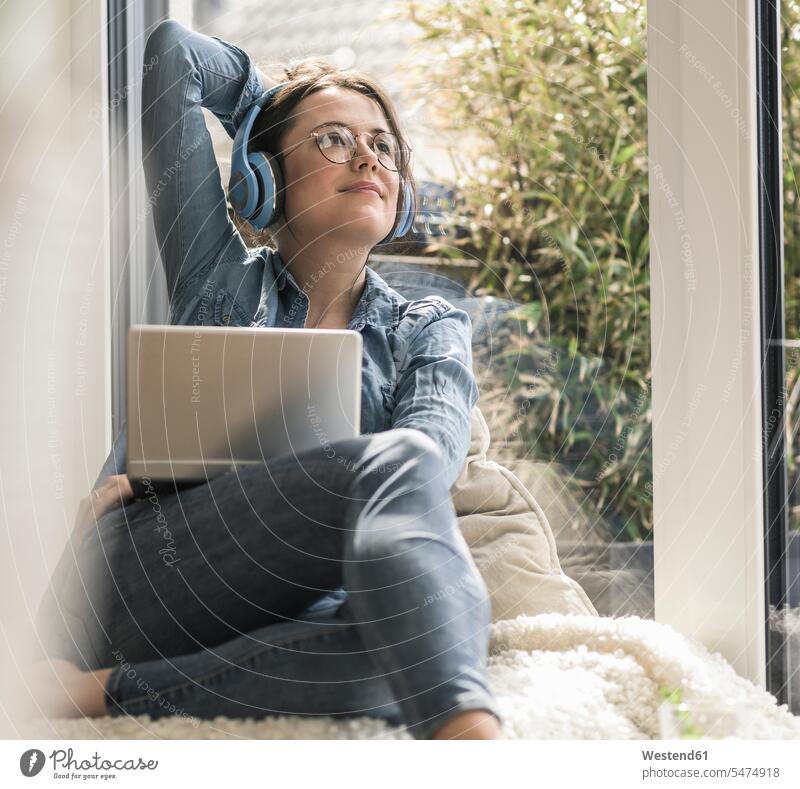 Woman with headphones and laptop sitting at the window at home headset windows woman females women smiling smile Laptop Computers laptops notebook Seated Adults