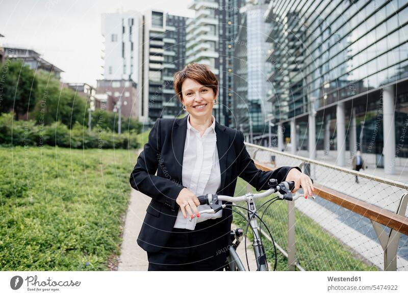 Smiling female professional with bicycle standing on footpath in city color image colour image outdoors location shots outdoor shot outdoor shots day