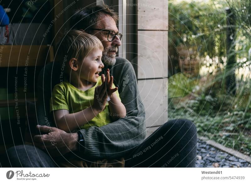 Father and little son sitting together on the floor at home looking out of window human human being human beings humans person persons families dad daddy