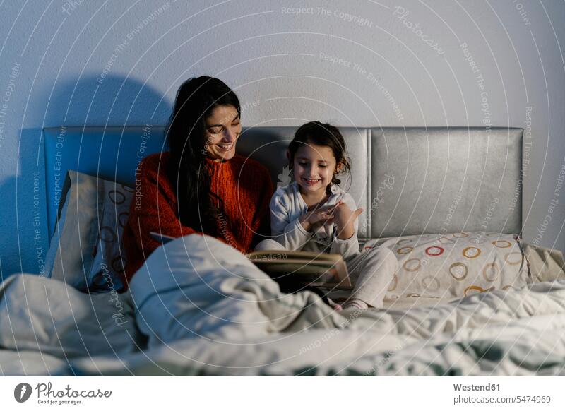 Happy mother and her little daughter sitting together on bed watching picture book human human being human beings humans person persons 2 2 people 2 persons two