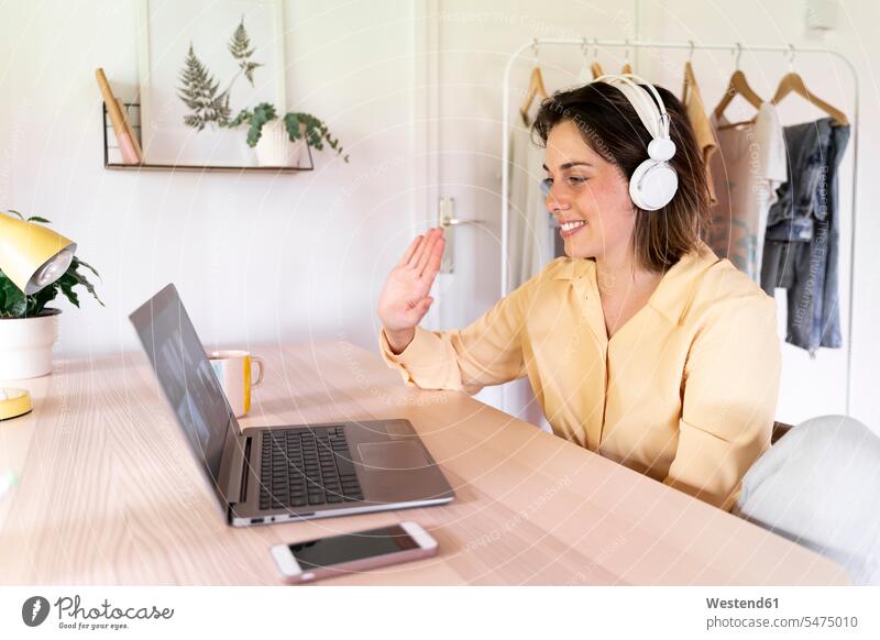 Smiling woman waving to friends on video call through laptop at home color image colour image Spain indoors indoor shot indoor shots interior interior view