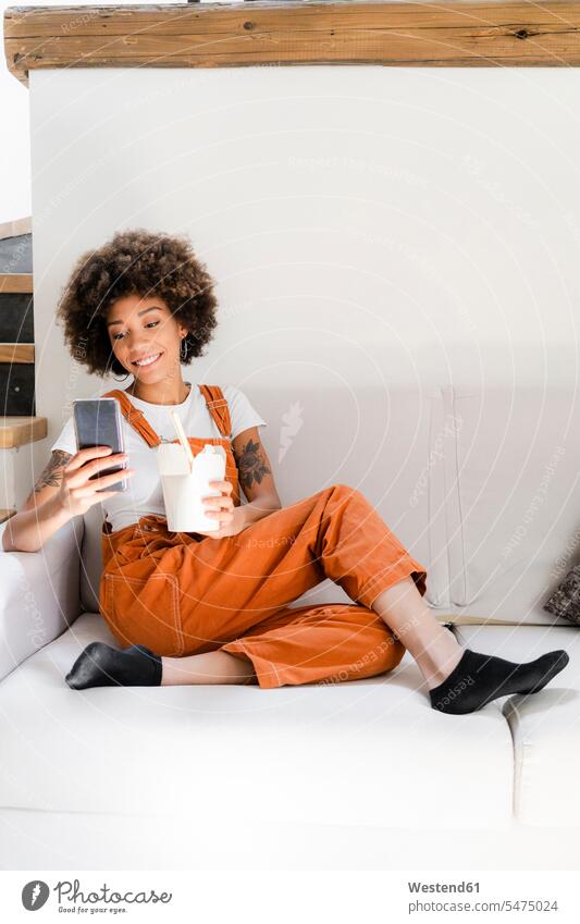 Portrait of smiling young woman sitting on the couch at home with box of Asian food looking at cell phone human human being human beings humans person persons