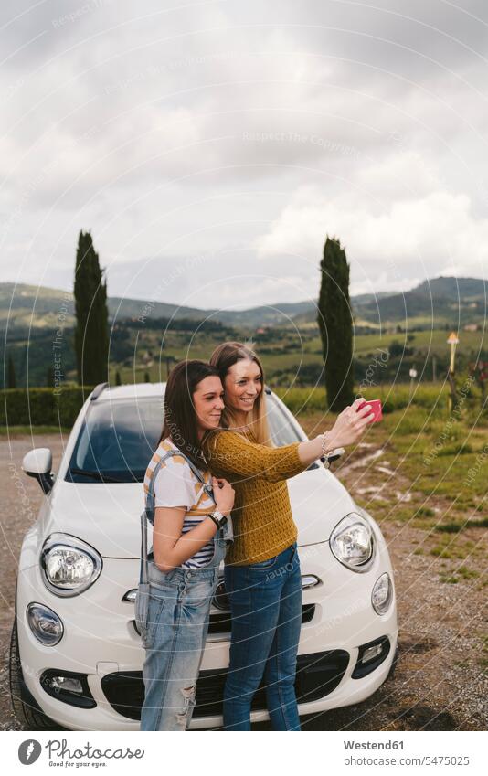 Two happy young women standing beside car taking a selfie, Tuscany, Italy human human being human beings humans person persons caucasian appearance