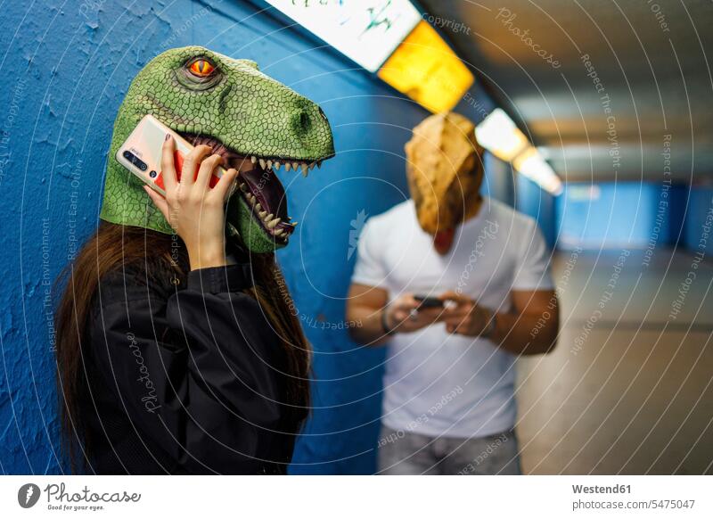 Female talking on smart phone by male friend while wearing dinosaur mask against blue wall color image colour image indoors indoor shot indoor shots interior