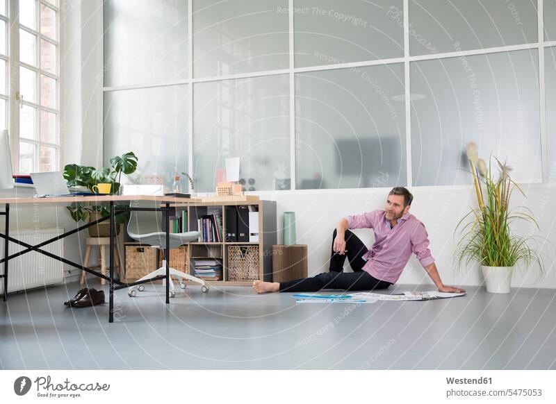 Casual man with plans sitting on the floor in a loft office lofts Seated casual men males offices office room office rooms floors Adults grown-ups grownups