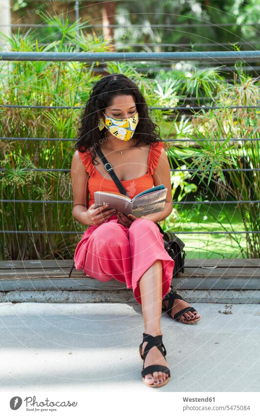 Young woman wearing protective mask while reading book sitting in park during COVID-19 pandemic color image colour image outdoors location shots outdoor shot