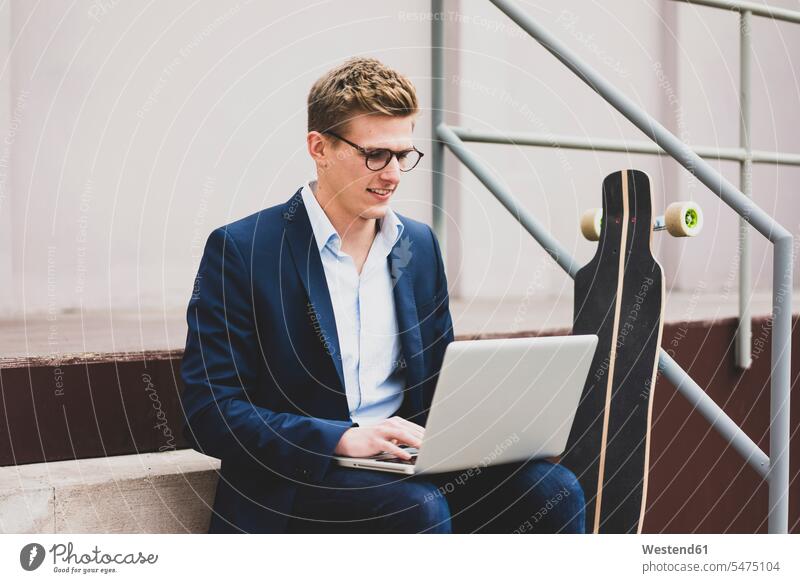Smiling young businessman with skateboard sitting outdoors on stairs using laptop smiling smile men males stairway Seated Businessman Business man Businessmen