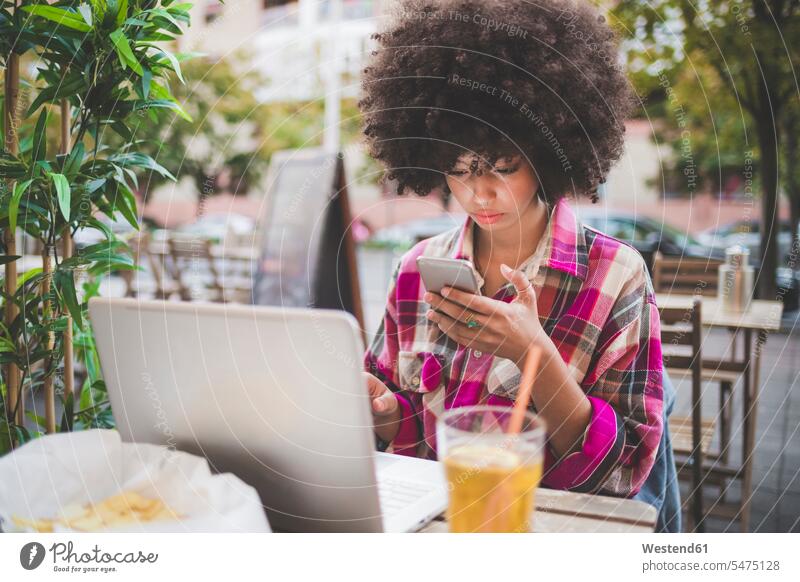 Young woman with afro hairdo using smartphone and laptop at an outdoor cafe in the city human human being human beings humans person persons curl curled curls