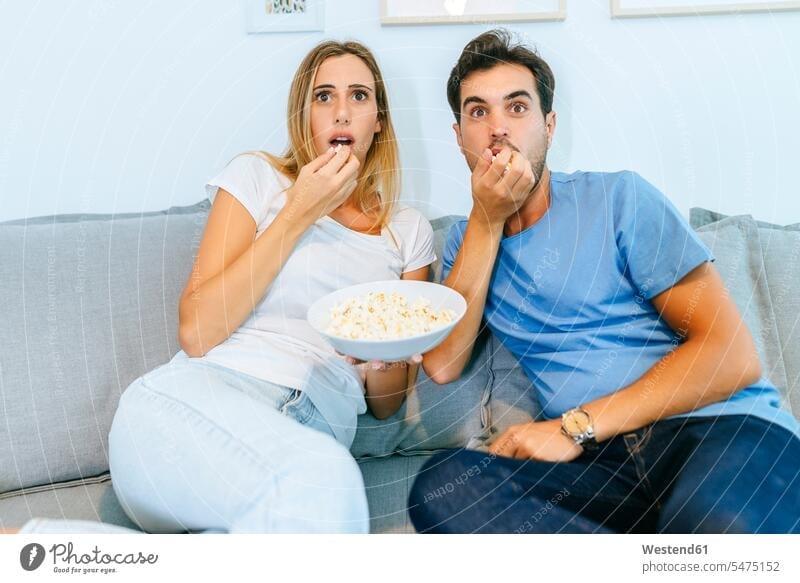 Excited couple watching TV while eating popcorn on sofa at home color image colour image indoors indoor shot indoor shots interior interior view Interiors day