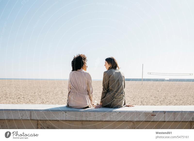 Back view of two friends sitting on the promenade enjoying leisure time female friends woman females women promenades free time indulgence enjoyment savoring