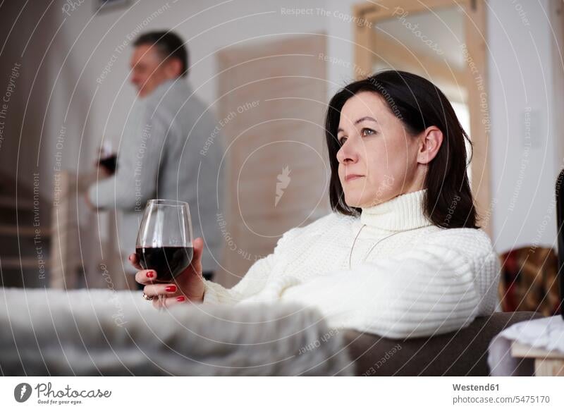 Serious mature woman with glass of wine and man in background Glass Drinking Glasses living room living rooms livingroom Red Wine Red Wines relaxed relaxation