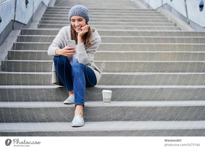 Happy woman with coffee to go sitting on stairs looking at cell phone Coffee females women happiness happy stairway Coffee to Go takeaway coffee Seated Drink