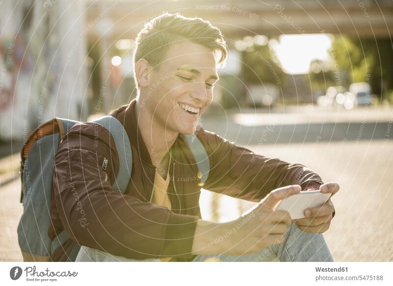 Happy young man using smart phone while sitting on street during sunny day color image colour image outdoors location shots outdoor shot outdoor shots