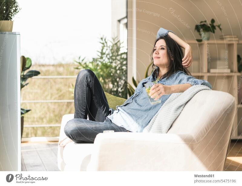 Smiling woman with a glass of infused water sitting on the couch at home females women Seated smiling smile Water settee sofa sofas couches settees Glass