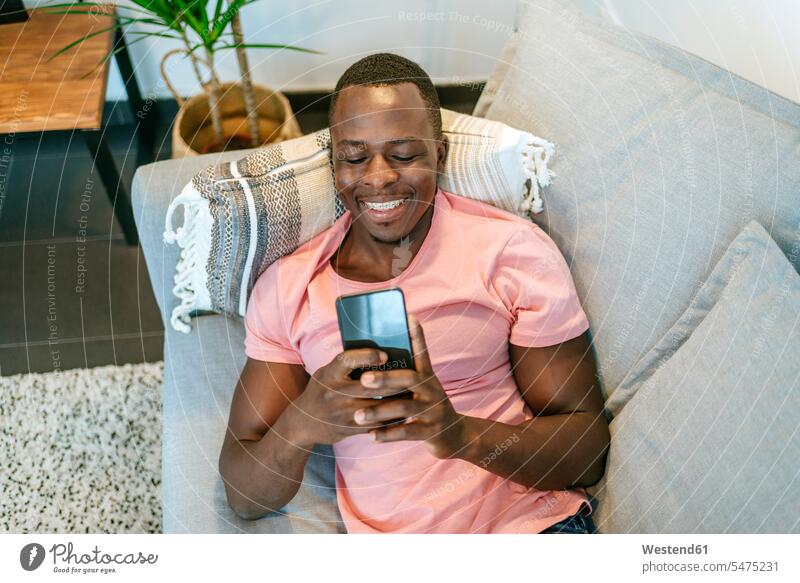 Happy young man lying on the couch with smartphone at home human human being human beings humans person persons African black black ethnicity coloured 1