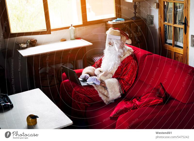 Man wearing Santa Claus costume using laptop while sitting on sofa at home color image colour image Spain leisure activity leisure activities free time