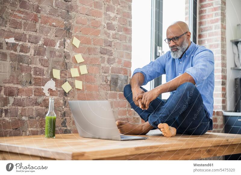 Businessman sitting on table in a loft using laptop Laptop Computers laptops notebook lofts Business man Businessmen Business men Seated use computer computers