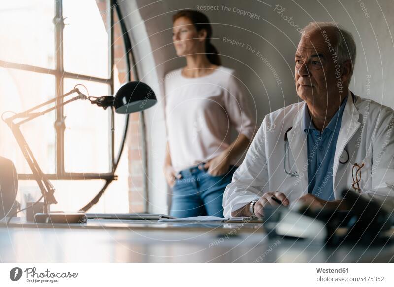 Serious senior doctor sitting at desk in medical practice with woman in background serious earnest Seriousness austere medical practices Doctors Office
