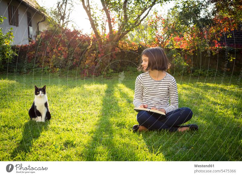 Smiling girl sitting on meadow with a book next to cat meadows females girls cats books smiling smile child children kid kids people persons human being humans