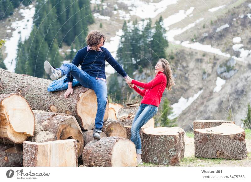 Cheerful woman pulling man sitting on log against mountain color image colour image outdoors location shots outdoor shot outdoor shots day daylight shot