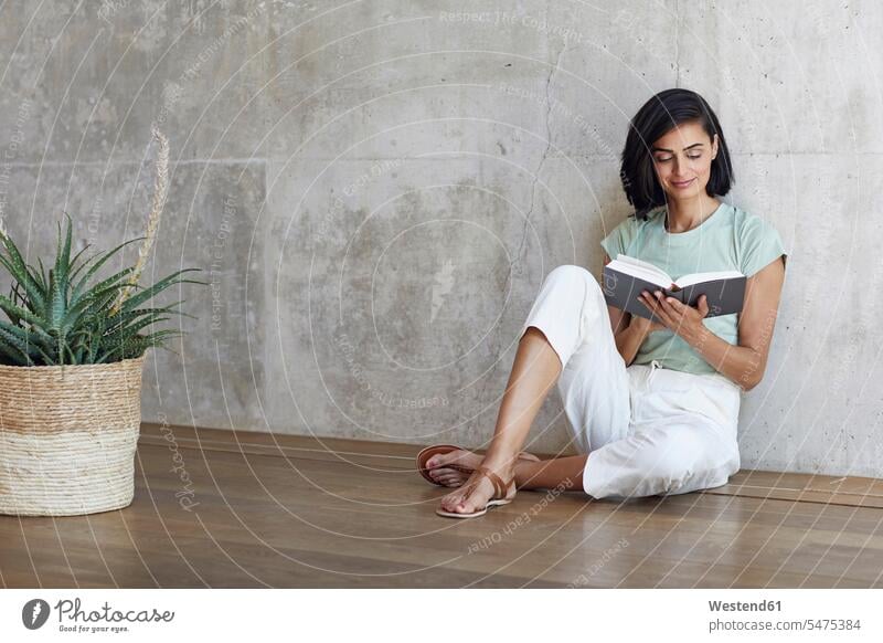 Businesswoman reading book while relaxing on hardwood floor against wall in office color image colour image Germany indoors indoor shot indoor shots interior