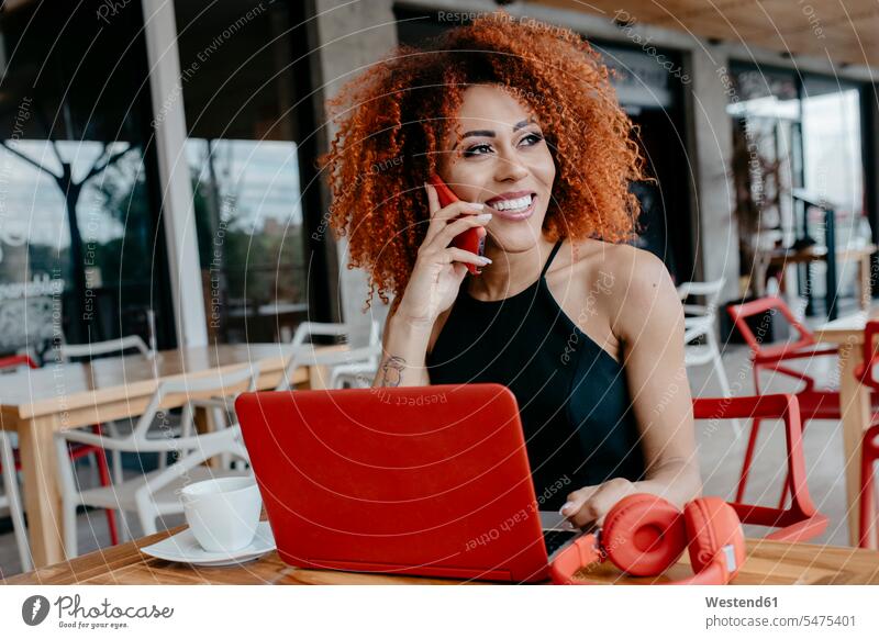 Afro woman with laptop and headphones on table talking over smart phone at sidewalk cafe color image colour image leisure activity leisure activities free time