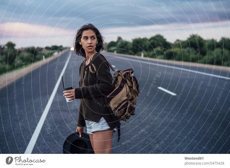 Portrait of young hitchhiking woman with backpack and beverage standing on lane rucksacks backpacks back-packs females women hitchhike hitch-hiking Lane Roadway